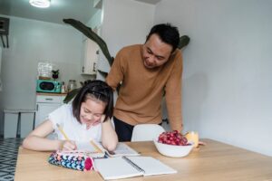 Asian father helping his daughter with homework at the kitchen table, fostering education and smiles.